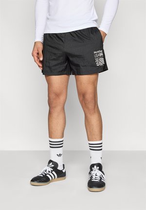 Schwarze Sportshorts mit strukturierter Oberfläche und weißem "World Cup USA 94"-Grafikdruck, kombiniert mit schwarzen Sneakers und weißen Socken mit Streifen.