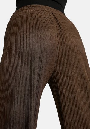 Hnědé široké kalhoty s texturou, které nosí člověk, se zaměřením na detail látky a střih kolem pasu a boků.