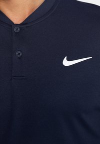 Marinblå pikétröja med en knappad krage och en vit Nike-logga på höger bröst. Tyget är slätt och lätt strukturerat.