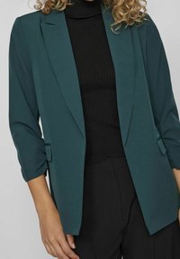 Blazer sarcelle à la coupe structurée, manches longues, revers crantés et poches latérales. Porté sur un col roulé noir côtelé.