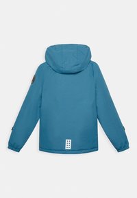 Blaue Kapuzenjacke mit glatter Textur, elastischen Bündchen und einem Logopatch auf der linken Schulter. Merkmale eine rechteckige Applikation mit Punkten auf der Rückseite.