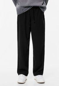 Osoba w czarnych spodniach dresowych na sznurki, białych trampek i szarym swetrze z prążkowanym wykończeniem, stojąca na tle białym.