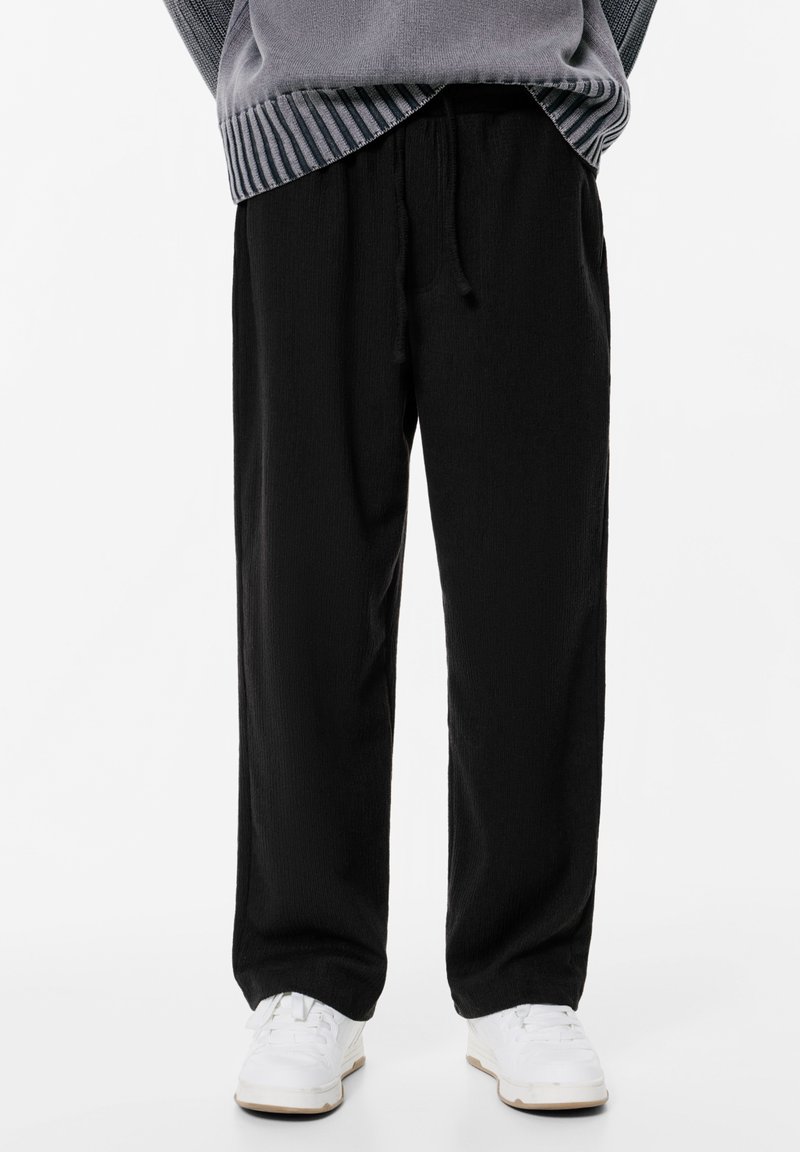 Osoba w czarnych spodniach dresowych na sznurki, białych trampek i szarym swetrze z prążkowanym wykończeniem, stojąca na tle białym.