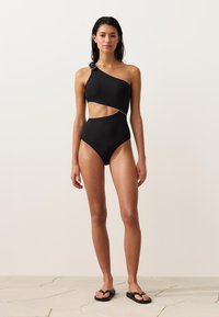 Femeie stând desculță pe podea de faianță, purtând un costum de baie întreg negru cu o singură bretea și decupaj, având un detaliu înnodătură pe bretea.