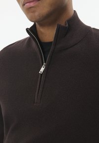 Man wearing dark brown textured quarter-zip sweater over a black shirt, with zipper partially open, against a white background.