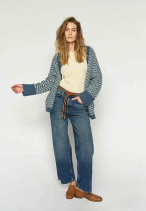 Frau trägt eine blau-weiß gestreifte Strickjacke, einen cremefarbenen Pullover, blaue Jeans mit einem braunen Gürtel und braune geflochtene Loafers, steht vor einem schlichten Hintergrund.