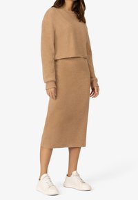 Ensemble en tricot beige composé d'un pull court et d'une jupe longue. La texture est douce, avec une coupe décontractée et des poignets côtelés, associé à des baskets blanches.