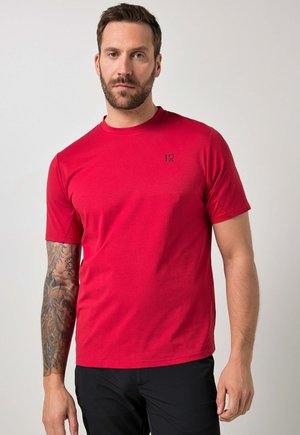 Mand med kort hår og skæg iført en rød T-shirt med kortærmer og sorte bukser, der viser en fuld tatoveringsærme på sin venstre arm.