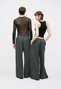 Schwarzes Tanktop und durchscheinendes schwarzes Langarmshirt kombiniert mit grauen, weit geschnittenen Jogginghosen mit roten Akzenten, von hinten gesehen.