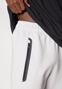 Γκρι αθλητικό σορτς με απαλή υφή, με ασύμμετρο μαύρο τσεπάκι με φερμουάρ. Ελαστική μέση και χαλαρή εφαρμογή.