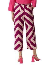 Spodnie z szerokimi nogawkami w kremowej bazie z wyrazistymi magentowymi wzorami w zygzak. Modelka nosi różowe sandały na obcasie, prezentując luźny krój.