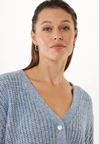 Kobieta o jasnej skórze i zielonych oczach, nosząca srebrne kolczyki w kształcie kół oraz niebieski sweter o fakturowanym splocie z białym guzikiem, na białym tle.