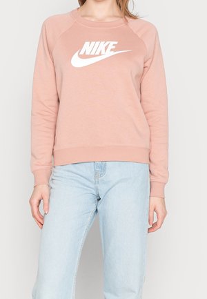 Kobieta w jasno różowej bluzie Nike z białym logo i jasnoniebieskich dżinsach, stojąca na jednolitym tle.