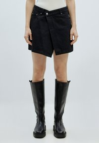 Schwarzer Denim-Wickelrock mit Vorderüberlappung, gesäumten Kanten und zwei vorderen Taschen. Kombiniert mit knielangen schwarzen Lederstiefeln.