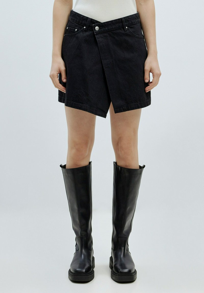 Schwarzer Denim-Wickelrock mit Vorderüberlappung, gesäumten Kanten und zwei vorderen Taschen. Kombiniert mit knielangen schwarzen Lederstiefeln.