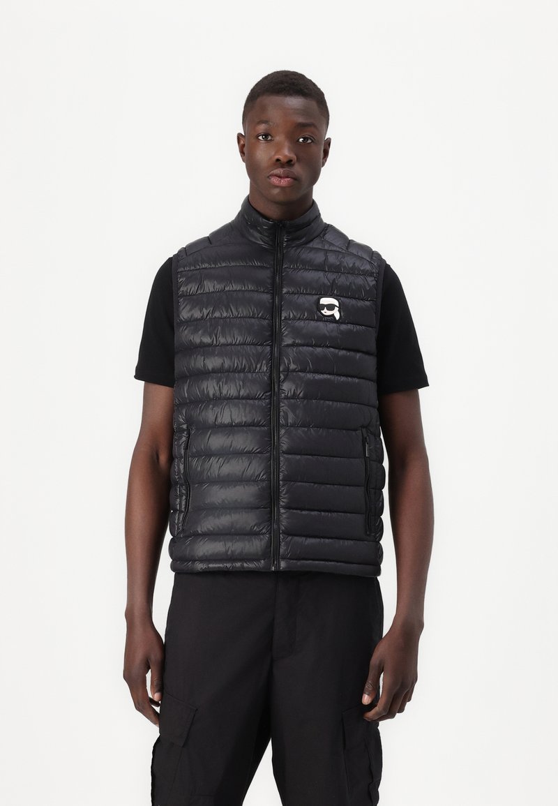 Melnas puffa veste ar horizontālu quilted dizainu, priekšējo rāvējslēdzēju un sānu kabatām. Iezīmēta ar mazu logotipu kreisajā krūtī.