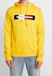 Hombre vestido con una sudadera amarilla brillante con bolsillo delantero, logo en bloque en negro, blanco y rojo que dice "Tommy Hilfiger", combinada con jeans de color azul claro.