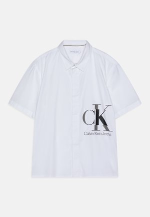 Chemise blanche à manches courtes avec un bouton, ornée du logo Calvin Klein Jeans en grandes lettres grises du côté droit, près du bas.