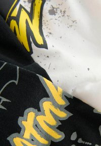 Grafisk t-shirt med gul och svart text på vit bakgrund, med en strukturerad yta och en blandning av djärva mönster.