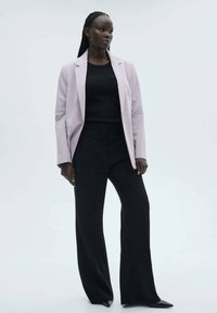 Hellrosa Blazer über einem schwarzen, figurschmeichelnden Top, kombiniert mit lockeren schwarzen Hosen. Glatter Stoff mit minimalen Details und maßgeschneiderter Passform.