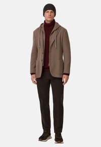 Brązowa kurtka z wełny z kapturem, bordowy golf, ciemne spodnie i brązowe trampki. Zawiera guziki i przednie kieszenie.