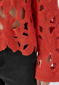 Czerwona dzianinowa sweter z kwiatowymi wycięciami, długimi rękawami i teksturowanym wykończeniem, w połączeniu z ciemnymi dopasowanymi spodniami.