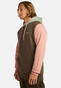Sudadera con capucha de cuerpo marrón, mangas rosas y capucha verde clara. Cuenta con un bolsillo frontal y un logo bordado en el pecho.