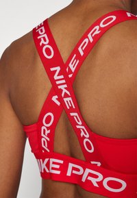 Sujetador deportivo rojo con tirantes en forma de cruz. Presenta el logotipo "NIKE PRO" en blanco en los tirantes. Textura suave y material elástico para soporte.