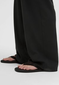 Pieds portant des tongs noires partiellement couverts par un long pantalon noir, debout sur une surface unie de couleur claire.
