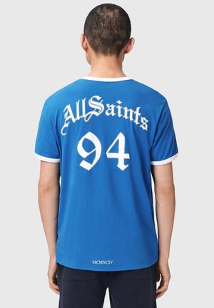 Mężczyzna noszący niebieski t-shirt AllSaints z białą lamówką, dużą cyfrą „94” i napisem „AllSaints” na plecach, stoi tyłem na jednolitym tle.