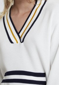 Vitt stickat tröja med V-ringning, med marinblå och gula ränder på kragen och fållen. Texturerad tyg med sportiga designelement.