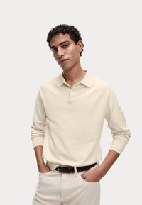 Dlouhý rukáv polo triko v off-white bavln�ěné směsi s límečkem. Žebrované manžety a uvolněný střih. Černý opasek zvýrazňuje světle barevné kalhoty.