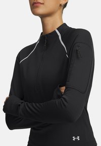 Persoon in een zwart sportshirt met lange mouwen en een ritskraag, reflecterende strepen op de schouders en een ritszak op de linker mouw.
