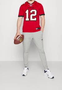 Punainen NFL-pelipaita, jossa mustat koristeet ja valkoinen numero 12, yhdistetty harmaisiin verkkareihin. Valkoiset lenkkitossut, joissa laivastonsiniset yksityiskohdat, ja kädessä ruskea jalkapallo.