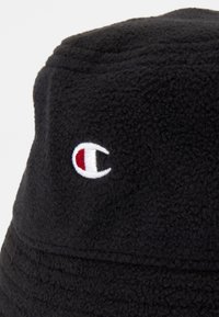 Μαύρο καπέλο bucket από φλις με λευκό και κόκκινο κεντητό λογότυπο στο πλάι. Μαλακό, υφασμάτινο υλικό με στρογγυλή κορυφή και φαρδύ γείσο.