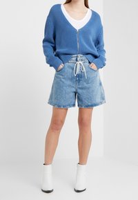 Ljusblå ribbad cardigan med dragkedja, bärs över en vit T-shirt. Kombineras med ljusa denimshorts, med dragsko i midjan och mjuk textur.