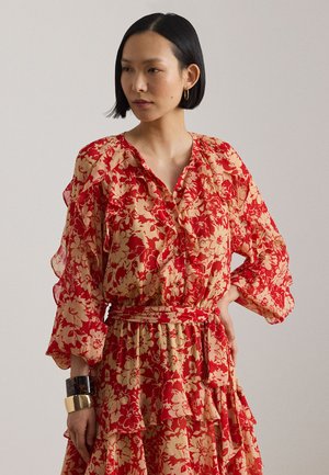 Mujer con cabello corto y negro, vestida con un vestido de manga larga rojo y beige con estampado floral, volantes y un lazo en la cintura, posando con una mano en la cadera.