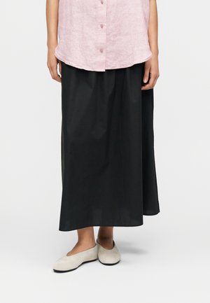 Ανοιχτό ροζ λινό πουκάμισο με κουμπιά σε συνδυασμό με μακρύ μαύρο φόρεμα. Η εμφάνιση ολοκληρώνεται με γκρι slip-on παπούτσια με μαλακή υφή.