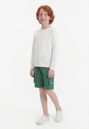 Smilende rødhåret barn står, iført hvit langermet skjorte, grønne cargoshorts og hvite slip-on sko mot hvit bakgrunn.