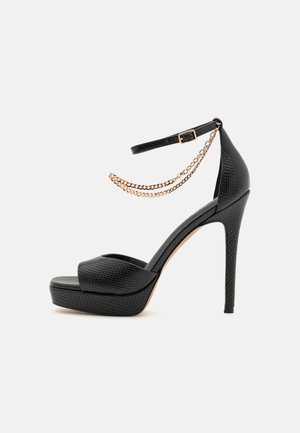 Czarna sandałka na wysokim obcasie z teksturowaną powierzchnią, platformą, paskiem wokół kostki oraz dwoma złotymi zdobieniami z łańcuszków na białym tle.