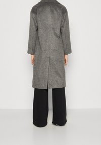 Persona vestida con un abrigo de lana gris de longitud media y pantalones negros de pierna ancha, de pie mirando hacia atrás contra un fondo claro y liso.