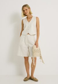 Top sans manches en mélange de lin blanc avec des rayures verticales, associé à un short assorti et un sac tissé avec des accents à franges. Les chaussures présentent des trous décoratifs.