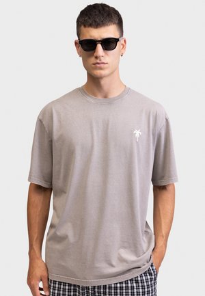 Άνδρας με γκρι μπλουζάκι που έχει μικρό λευκό λογότυπο φοίνικα, μαύρα γυαλιά ηλίου και καρό σορτς, στέκεται μπροστά σε ένα απλό ανοιχτόχρωμο φόντο.