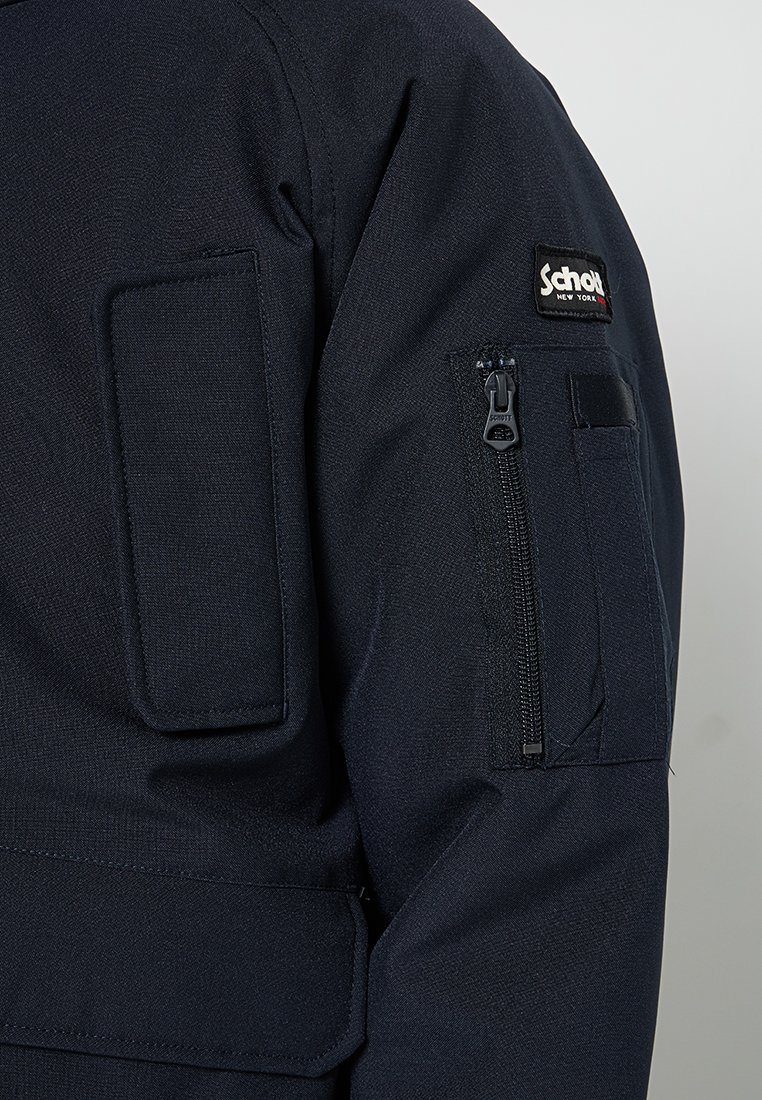 Vêtement d'extérieur bleu marine en tissu texturé, avec une grande poche plaquée et une poche zippée sur la manche. Étiquette de marque visible.