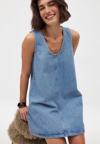 Denim-mekko, jossa on hihaton malli, pyöreä kaula-aukko, sininen väri ja näkyvät ompeluyksityiskohdat. Kankaan pinta näyttää olevan sileä ja malli on rento.