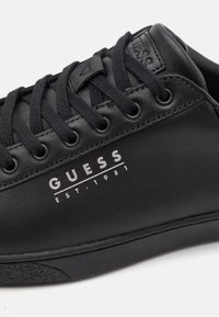 Must nadava musta madal poolsaapad tekstureeritud nahast pealsetega, mustade paeltega ja ümmarguste silmadega. Küljel on valge "GUESS EST.1981" logo.