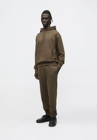 Καφέ φούτερ με κουκούλα και αντίστοιχο παντελόνι, με μπλε λογότυπο, κατασκευασμένα από απαλό ύφασμα, με άνετη εφαρμογή και λεπτομέρειες με κορδόνια.