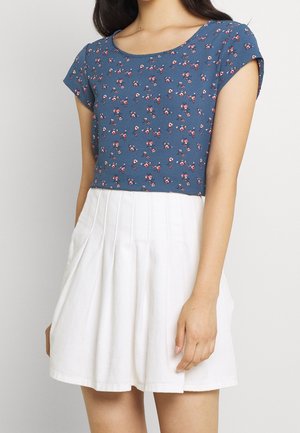 Vrouw met een blauw bloemenprint shirt met korte mouwen, gecombineerd met een witte geplooide rok, staand tegen een effen lichte achtergrond.