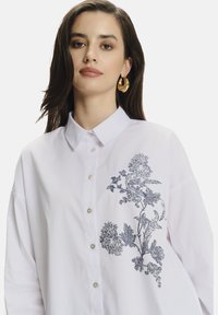 Camisa blanca con cuello abotonado, que presenta bordados florales azules en el lado izquierdo. Tejido de algodón suave con un corte relajado.