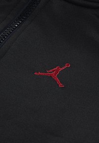 Black fabric with a subtle texture features a red embroidered Jumpman logo, positioned near a smooth zipper.